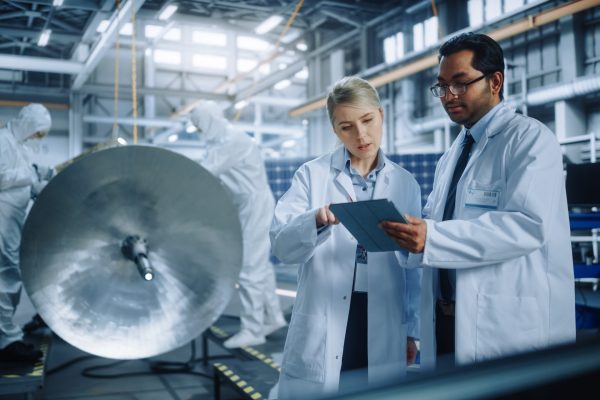 Team of Engineers Working on Satellite Construction, Use Tablet Computer Talk about Data. Aerospace Spaceship Manufacturing Facility: Team of Scientists Building Spacecraft for Space Exploration